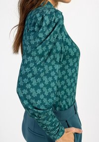 Blusa verde acqua con motivo floreale texturizzato, maniche a sbuffo e polsini arricciati. Indossata con pantaloni abbinati a vita alta.