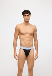 Crni muški tanga s elastičnom trakom svijetlo plave boje na kojoj se nalazi logo "Calvin Klein", izrađen od rastezljivog materijala za udobnost.