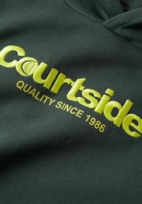 Zelená mikina s kapucí z texturované látky, s výrazným žlutým vyšitým nápisem "courtside QUALITY SINCE 1986."