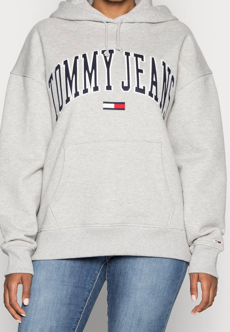 Personne portant un sweat à capuche Tommy Jeans gris clair avec poche avant et jean bleu, affichant le logo de la marque et l'emblème du drapeau sur la poitrine.