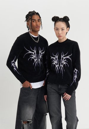 Svart strikkegenser med lilla og hvite lynmønstre, båret av to modeller med mørke jeans, som står side om side.