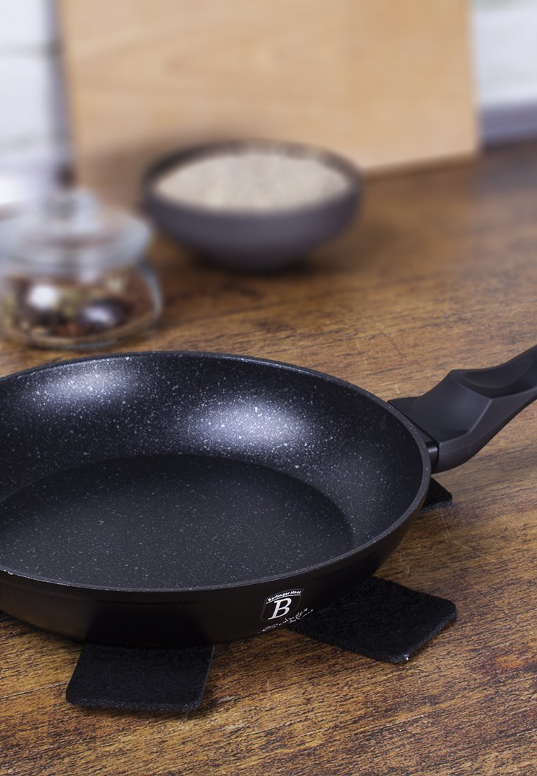 Schwarze Antihaft-Bratpfanne mit glatter Oberfläche, flachem Boden und ergonomischem Kunststoffgriff. Auf schwarzen Untersetzern auf einer Holzoberfläche platziert.