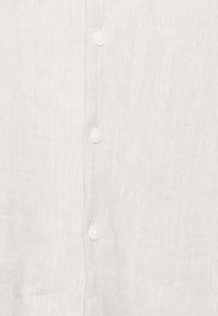 Chemise en lin beige clair avec une fermeture à boutons sur le devant, dotée de boutons blancs. Le tissu a une apparence douce et texturée.