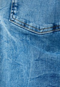 Tela de mezclilla en azul claro. Primer plano de un bolsillo trasero con forma rectangular. Presenta costuras rectas y una superficie texturizada.