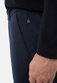 Granatowe spodnie o gładkiej fakturze, z boczną kieszenią i małym srebrnym logo B. Widoczne ciemne mankiety swetra nad nimi.