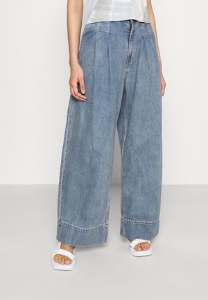 Wijde blauwe denim jeans met plooien aan de voorkant, gedragen met witte platformsandalen en een wit topje, getoond van taille tot voeten.