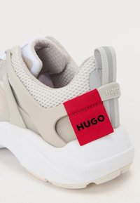 Hellgrau Sneaker mit einem strukturierten Mesh-Obermaterial, Kunststoffakzenten, einem roten Stoffetikett mit "HUGO" und einer weißen, klobigen Sohle.