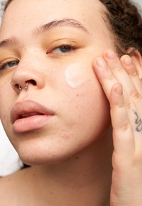 Person som applicerar en kräm på sin kind; huden är ljus med fräknar. Synlig septum-piercing, ljusögd och texturerad hår.