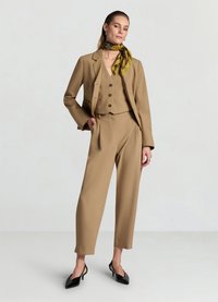 Beiger maßgeschneiderter Anzug bestehend aus einer taillierten Jacke und einer hochgeschnittenen Hose, kombiniert mit einem gemusterten grünen Schal und schwarzen spitzen Schuhen.