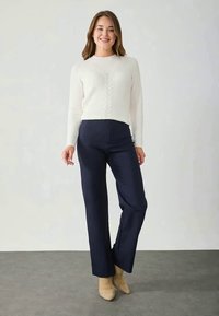 Pulover alb cu model côtelat și tricotare în cablu, asortat cu pantaloni largi bleumarin. Modelul poartă botine bej. Ansamblu simplu, în nuanțe neutre.