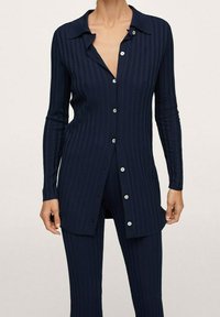 Žena oblečená v tmavě modré žebrované košili s dlouhým rukávem a zapínáním na knoflíky a sladěných kalhotách, nafocená od krku po polovinu stehna.