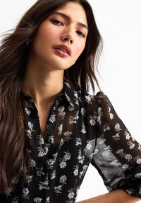 Black sheer blouse with floral prints, button-up design, and collar; features long sleeves and a lightweight texture.
