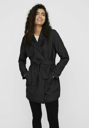 Manteau noir en trench avec une taille ceinturée, des boutons double boutonnage, des poches latérales et une texture lisse. Porté par-dessus un haut clair et un pantalon noir.