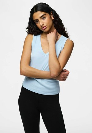Young woman with long curly black hair wearing a light blue sleeveless top and black pants, posing with one hand on her neck and the other crossed.