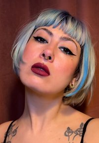 Coiffure bob courte avec des tons bleu clair et argenté. Eyeliner audacieux en aile, lèvres rouge foncé, multiples piercings faciaux et tatouages floraux visibles.