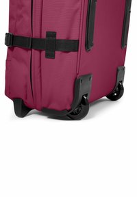 Röd mjuk resväska med svarta hårdplastdetaljer, två hjul och ett säkerhetsband. Texturerad yta och förstärkta hörn.