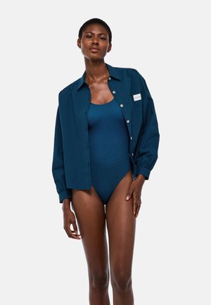 Femme à la peau foncée portant un maillot de bain côtelé bleu foncé et une chemise assortie déboutonnée, sur un fond clair uni.