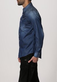 Jeansskjorta i mörkblått. Har en knappstängning fram, bröstfickor och långa ärmar med knäppning vid handlederna. Slät textur med en lätt lyster.