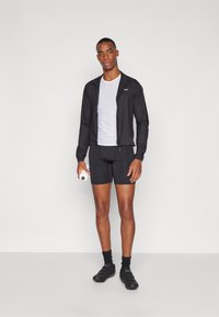 Idrottsoutfit bestående av en lätt svart zip-jacka, en vit mesh-tröja, svarta shorts och svarta cykelskor, med en vattenflaska i handen.