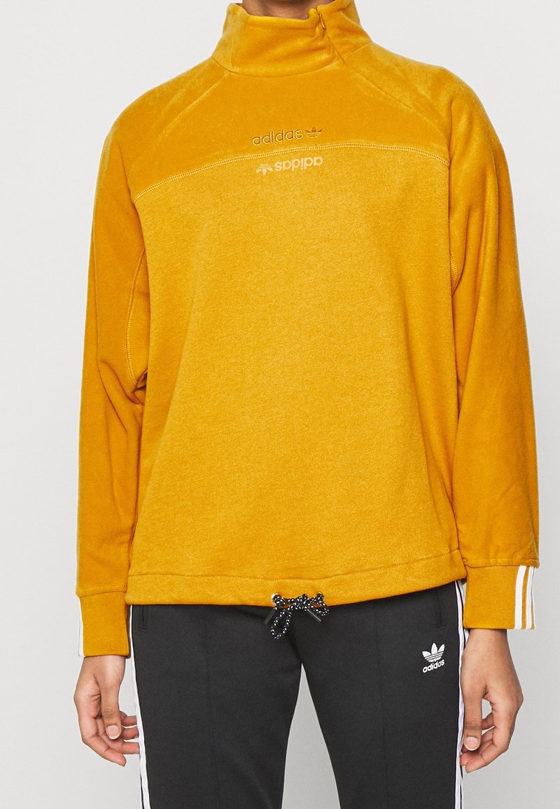 Gelber Fleece-Pullover mit hohem Kragen, der einen halben Reißverschluss an der Vorderseite hat und drei Streifenakzente an den Ärmeln aufweist. Schwarze Kordel am Saum.