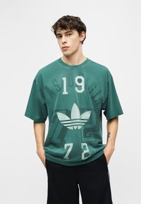 Jeune homme aux cheveux bouclés portant un t-shirt vert ample Adidas avec des chiffres blancs et un pantalon noir, les mains dans les poches, expression neutre.