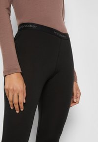 Zwarte leggings met een gladde textuur en een stretchy tailleband waarop de merknaam "icebreaker" in het grijs staat. De hand van het model rust op de dij.