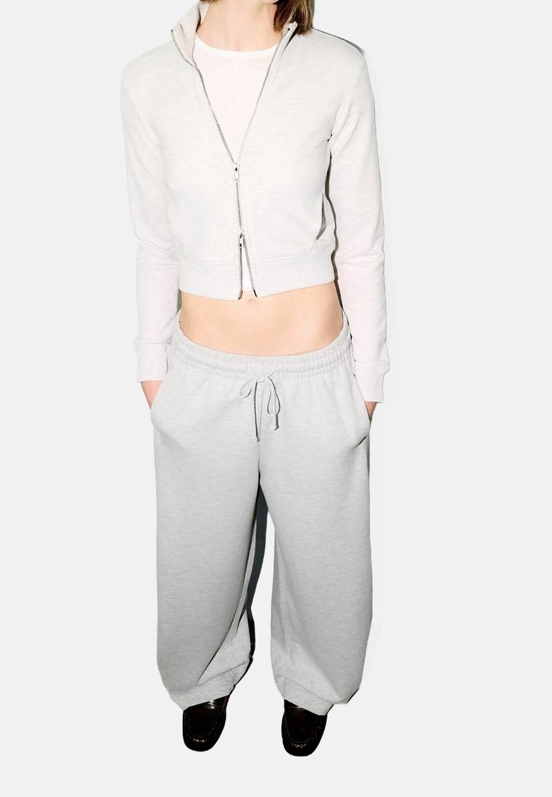 Ljusgrå kort zip-up-jacka och matchande mjukisbyxor med vida ben; mjukt tyg, synlig dragsko och sidofickor.