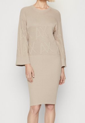 Beżowa sukienka dziana z teksturowaną górą i ołówkową spódnicą z prążkowanego materiału, z długimi rękawami i okrągłym dekoltem, noszona przez stojącą osobę na jednolitym tle.