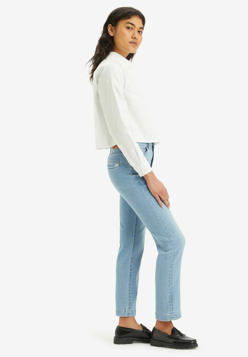 Gekürzte weiße Hemdbluse, hellblaue schmale Jeans mit leichtem Stretch und schwarze Slipper. Das Model steht seitlich vor einem weißen Hintergrund.