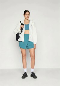 Junge Frau in teal Sport-Bra und Shorts, weißem Reißverschluss-Hoodie, schwarzen Sneakers, weißen Socken und mit einem schwarzen Rucksack.