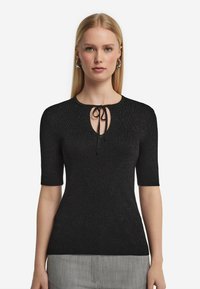 Svart stickad topp med korta ärmar, med rund ringning och en nyckelhålsöppning knuten med en svart dragsnodd. Subtil glittrande struktur.