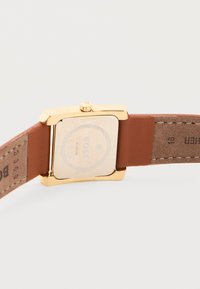 Χρυσό ρολόι χειρός με τετράγωνο καντράν και καφέ δερμάτινο λουράκι, με την πλευρά της αγκράφας προς τα κάτω πάνω σε λευκή επιφάνεια, δείχνοντας την πίσω όψη του ρολογιού και τις λεπτομέρειες των ραφών.