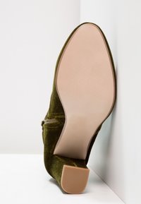 Bottines en velours vert olive avec semelle beige et talon carré, positionnées debout contre un mur blanc en coin.