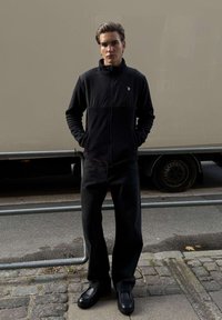 Mladý muž v čiernej bunde na zips, čiernych nohaviciach a čiernych topánkach stojí s rukami v vreckách na dláždenej ulici pri kovovom zábradlí.