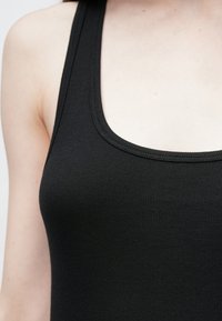 Krupni plan gornjeg dijela tijela u crnoj rebrastoj majici bez rukava s širokim naramenicama i zaobljenim izrezom na blijedoj koži.