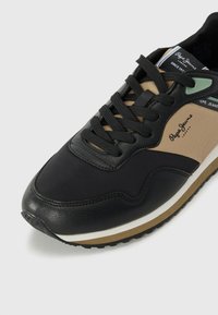 Černé tenisky se směsí syntetických tkanin a kůže. Mají béžovou část, černé tkaničky, bílou mezipodešev a zelený akcent u ok. na tkaničky.