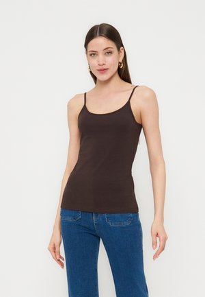 Frau mit langen braunen Haaren, die goldene Creolen, ein eng anliegendes dunkelbraunes ärmelloses Oberteil und blaue Jeans trägt, steht vor weißem Hintergrund.