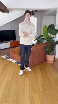 Ανοιχτό μπεζ πουλόβερ με φερμουάρ μέχρι το ένα τέταρτο, σκούρα τζιν και άσπρα αθλητικά παπούτσια. Στο φόντο υπάρχει καφέ καναπές με κορδόνια, γλάστρα με φυτό και ξύλινο πάτωμα.