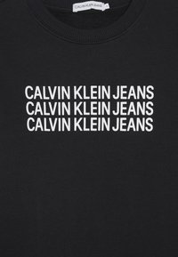 Czarna bluza z ribbowym dekoltem, na której z przodu widnieje powtarzający się napis "CALVIN KLEIN JEANS" w białych, pogrubionych, wielkich literach.