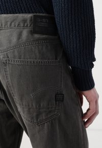 Tamno sive G-Star Raw traperice, s teksturiranim materijalom, stražnjim džepovima i crnom kožnom oznakom na pojasu s utisnutim logom.