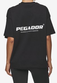 Person wearing a black oversized T-shirt with white "PEGADOR" text and small print "PEGADOR ALL RIGHTS RESERVED" on the back.