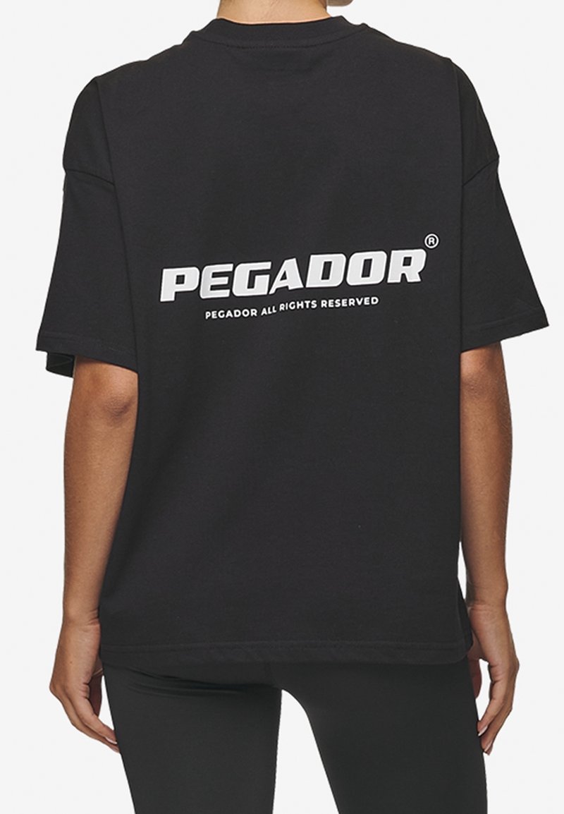 Person wearing a black oversized T-shirt with white "PEGADOR" text and small print "PEGADOR ALL RIGHTS RESERVED" on the back.