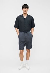 Νεαρός άνδρας που στέκεται με τα χέρια στις τσέπες, φοράει σκούρο πουκάμισο με κοντά μανίκια και κουμπιά, γκρι σορτς με κορδόνι, λευκές κάλτσες και λευκά αθλητικά παπούτσια.