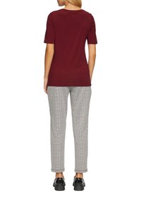 Vrouw die met de rug naar ons toe staat, gekleed in een bordeauxrood T-shirt met korte mouwen, geruite broeken en zwarte instappers op een witte achtergrond.