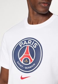 Hvid bomulds t-shirt med et rundt Paris Saint-Germain-logo i marineblå og rød med en gul fleur-de-lis, pyntet med en rød Nike swoosh.
