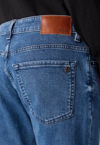 Vue de dos d'un jean en denim bleu avec un patch en cuir marron étiqueté BOSS au-dessus de la poche arrière droite, porté avec un haut noir.
