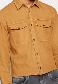 Koszula khaki z miękkiej tkaniny, z dwoma kieszeniami na piersi, zapinana na guziki z przodu, z ciemnymi guzikami. Charakteryzuje się casualowym krojem i widocznymi przeszyciami.