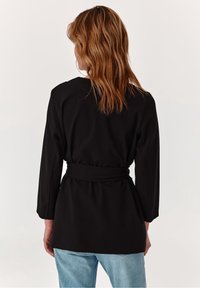 Svart bältejacka med långa ärmar, strukturerad tyg och en figursydd midja. Bakpanelen har en slät yta och en något sänkt fåll.