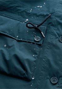 Tmavě tyrkysová voděodolná tkanina bundy s kapkami vody, která má knoflíkové zapínání, stahovací šňůrku a strukturovaný detail přední kapsy.