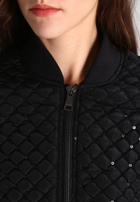 Czarna pikowana kurtka bomberka z prążkowanym kołnierzem, z suwakiem z przodu i subtelnymi cekinami podkreślającymi diamentowy wzór.